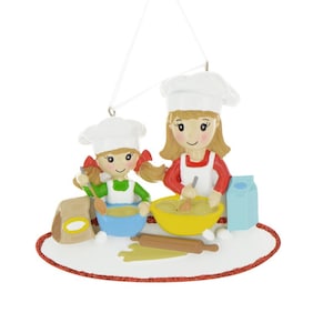May include: A white ceramic Christmas ornament depicting two people wearing chef hats and aprons, baking together. The ornament features a red and white striped apron, a green and white striped apron, a blue bowl, a yellow bowl, a rolling pin, and a bag of flour.
