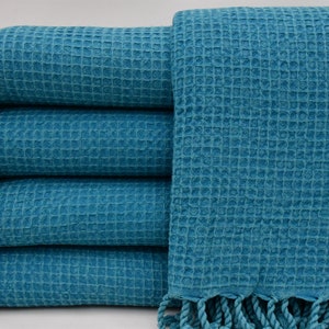 May include: A stack of five teal waffle weave towels with fringe trim. The towels are folded and stacked on a white surface.
