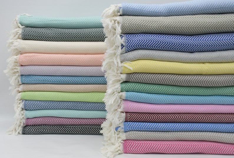 May include: A stack of colorful woven cotton throws with a herringbone pattern. The throws are folded and stacked in a variety of colors, including pink, blue, green, and yellow.