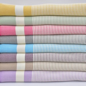 K&ouml;nnte beinhalten: Ein Stapel von acht gestreiften Handt&uuml;chern in verschiedenen Farben, darunter Gelb, Gr&uuml;n, Rosa, Grau, Blau, Lila, Beige und Lavendel. Die Handt&uuml;cher sind ordentlich gefaltet und gestapelt, wobei die Fransen an den Enden unten sichtbar sind.