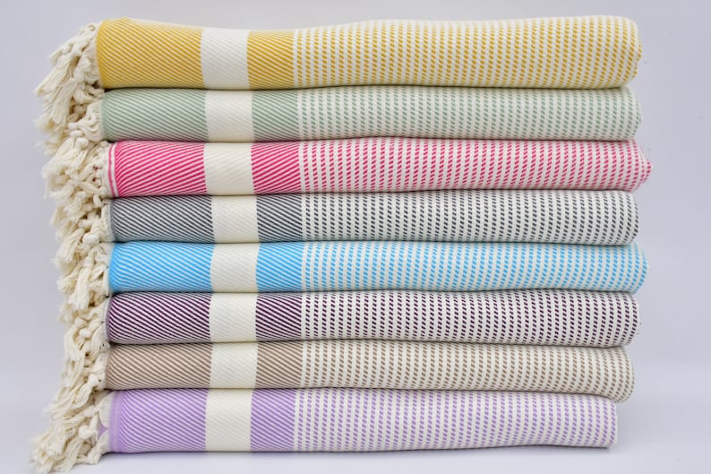 K&ouml;nnte beinhalten: Ein Stapel von acht gestreiften Handt&uuml;chern in verschiedenen Farben, darunter Gelb, Gr&uuml;n, Rosa, Grau, Blau, Lila, Beige und Lavendel. Die Handt&uuml;cher sind ordentlich gefaltet und gestapelt, wobei die Fransen an den Enden unten sichtbar sind.