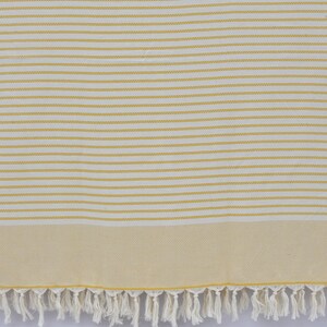May include: A white and yellow striped beach towel with a fringe border. The towel is made of cotton and is soft and absorbent.