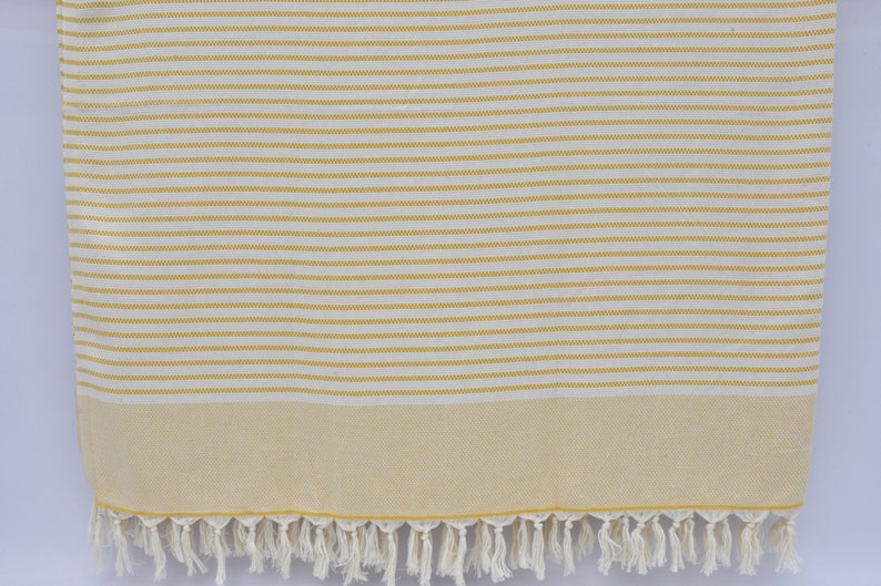 May include: A white and yellow striped beach towel with a fringe border. The towel is made of cotton and is soft and absorbent.