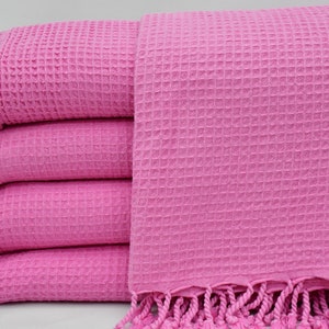 May include: A stack of four pink waffle weave towels with fringe trim. The towels are folded and stacked on a white surface.
