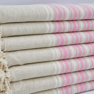 Può includere: Una pila di asciugamani beige a righe rosa e bianche. Gli asciugamani hanno un bordo con frange su un lato. Gli asciugamani sono piegati e impilati ordinatamente, mostrando il motivo a righe.