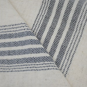 May include: White fabric with blue stripes woven into the material. The fabric is folded in a V-shape, showing the stripes from two different angles.