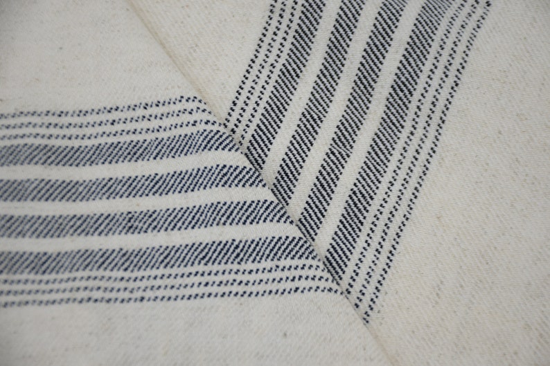 May include: White fabric with blue stripes woven into the material. The fabric is folded in a V-shape, showing the stripes from two different angles.