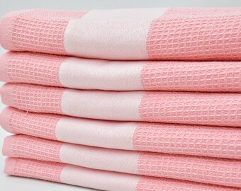 Può includere: Un mucchio di sei asciugamani rosa a nido d'ape con strisce bianche. Gli asciugamani sono piegati e impilati ordinatamente.