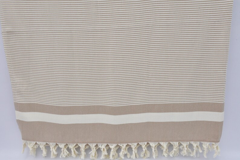 K&ouml;nnte beinhalten: Ein beige-wei&szlig; gestreiftes t&uuml;rkisches Handtuch mit Fransenrand. Das Handtuch hat eine dezente gewebte Textur.