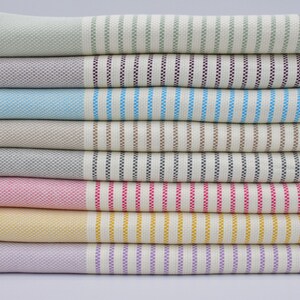 May include: A stack of eight striped towels in various colors, including green, purple, blue, brown, gray, pink, yellow, and lavender. The towels have a woven texture and fringe edges.