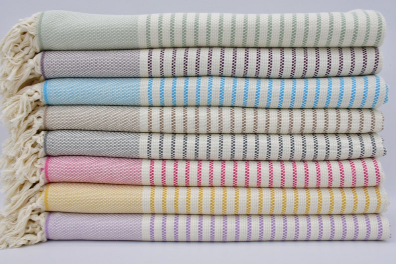 May include: A stack of eight striped towels in various colors, including green, purple, blue, brown, gray, pink, yellow, and lavender. The towels have a woven texture and fringe edges.