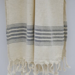 May include: A white cotton towel with blue stripes and fringe. The towel is folded in half and hanging on a white wall.