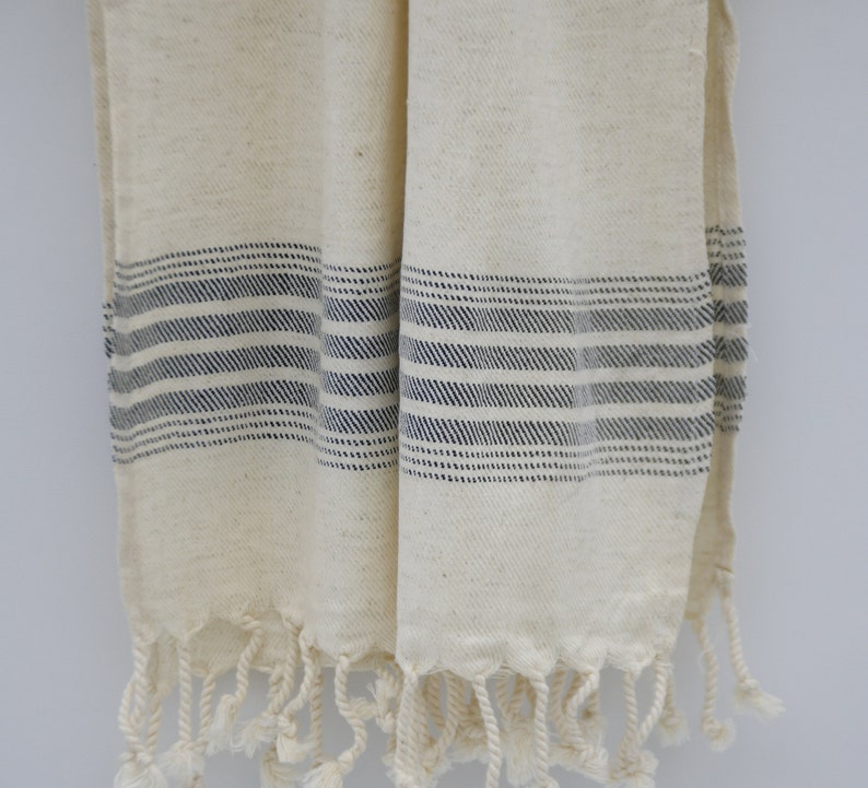 May include: A white cotton towel with blue stripes and fringe. The towel is folded in half and hanging on a white wall.