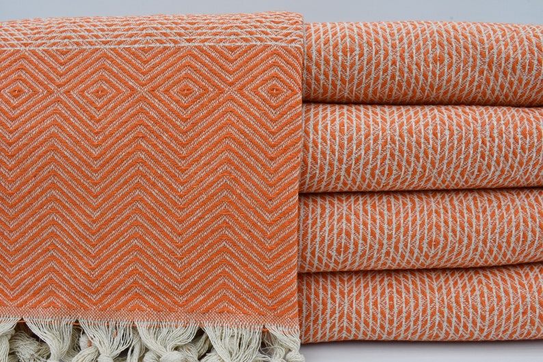 May include: A stack of orange and white patterned Turkish towels with fringe. The towels are made of cotton and are soft and absorbent.