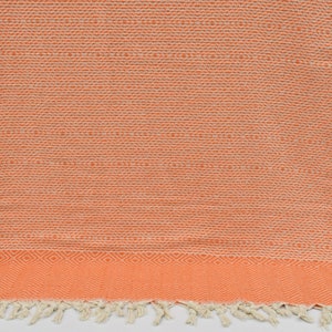 May include: A coral orange Turkish towel with a white fringe and a geometric pattern. The towel is made of cotton and is soft and absorbent.