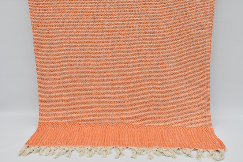 May include: A coral orange Turkish towel with a white fringe and a geometric pattern. The towel is made of cotton and is soft and absorbent.