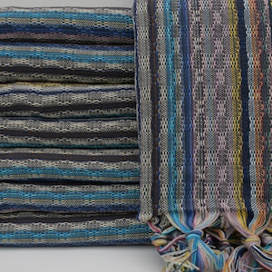 May include: A stack of woven Turkish towels with colorful fringe. The towels feature a striped pattern in shades of blue, gray, and white. The fringe includes yellow, pink, and blue threads.