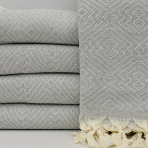 May include: A stack of four gray Turkish towels with a diamond pattern and white fringe. The towels are folded and stacked on a white surface.