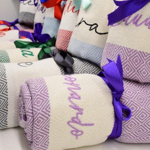 May include: A collection of rolled up towels with purple, green and grey stripes. Each towel has a different name embroidered on it in white thread. The towels are tied with purple and blue ribbons.
