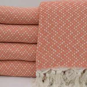 May include: A stack of four coral-colored woven cotton towels with a white geometric pattern. The towels have a fringed edge in a cream color. The towels are folded and stacked neatly, showcasing the texture and design.