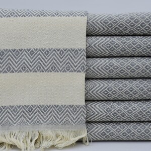 Puede incluir: Una pila de toallas estampadas grises y blancas. Las toallas presentan un diseño geométrico de diamantes y galones. Las toallas dobladas están apiladas ordenadamente, con un borde con flecos visible en la toalla superior.