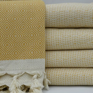 May include: A stack of yellow and white patterned Turkish towels with fringed edges. The towels are folded and stacked on a white surface.