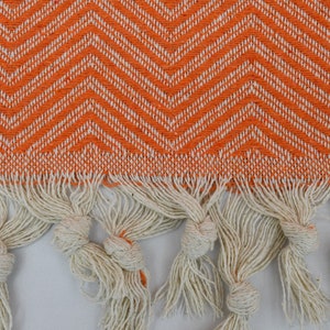May include: Orange and white chevron patterned fabric with a fringe border. The fringe is made of white cotton and is knotted at the ends.