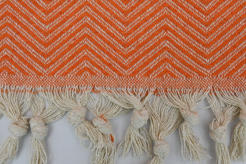 May include: Orange and white chevron patterned fabric with a fringe border. The fringe is made of white cotton and is knotted at the ends.