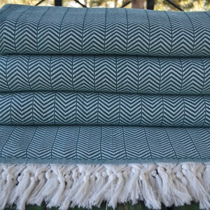 May include: A teal blue throw blanket with a white herringbone pattern and white fringe. The blanket is folded in fourths and is sitting on a wooden surface.
