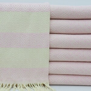 Puede incluir: Una pila de toallas rayadas de color rosa y blanco con flecos en la parte inferior. Las toallas están dobladas y apiladas sobre una superficie blanca.