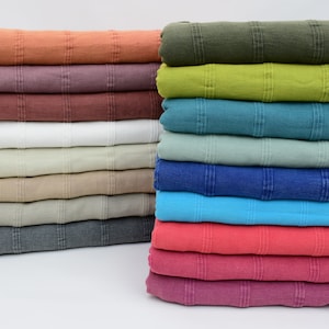 Può includere: Una pila di 17 coperte in cotone tessute di diversi colori con bordi a frangia. Le coperte sono disposte in una pila verticale, con la coperta superiore di colore verde scuro e la coperta inferiore di colore viola scuro.