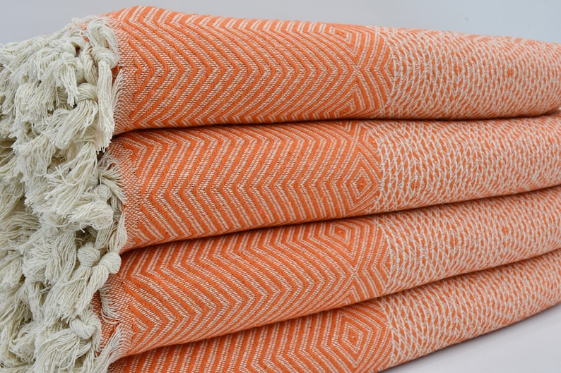 May include: A stack of orange and white patterned Turkish towels with fringe. The towels are folded and have a chevron pattern.