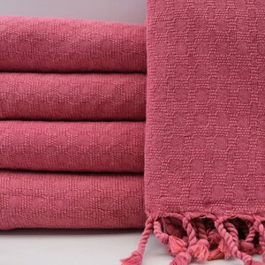 May include: A stack of five pink woven towels with fringe. The towels are folded and stacked on a white surface.