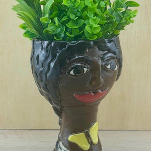 May include: A decorative ceramic planter shaped like a face, with dark brown skin, black hair, and red lips. The planter features a yellow butterfly accent and is filled with green artificial plants. The base has a white and black pattern.