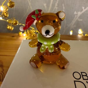 May include: A glass bear figurine wearing a Christmas hat and a green scarf. The bear is brown with white details on its ears and face. The hat is red with white trim and holly berries. The bear is on a white surface.