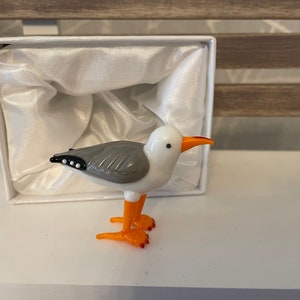 May include: A small glass seagull figurine with white and grey feathers, orange legs and beak, and black eyes. The figurine is in a white box with a white satin lining.