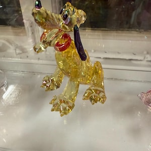 May include: A yellow glass dog figurine with a red collar and purple ears. The dog is in a sitting position with its front paws extended. The glass has an iridescent, reflective quality.