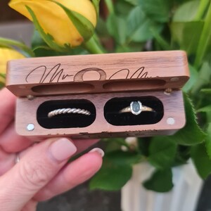 May include: A wooden ring box with two compartments. The top compartment contains a silver band with a braided design. The bottom compartment contains a gold band with a black stone. The box is engraved with the words "Mr. & Mrs."