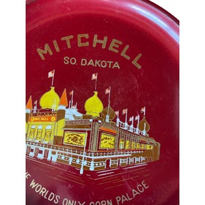 May include: A red metal tray with gold lettering and a detailed illustration of the Corn Palace in Mitchell, South Dakota. The text reads "MITCHELL SO. DAKOTA" and "THE WORLD'S ONLY CORN PALACE". The illustration features the building with flags.
