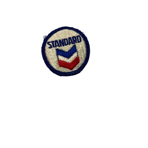May include: A circular patch with a blue border and the word "STANDARD" in blue lettering. Inside the circle is a white background with a blue, white, and red chevron design. The patch is likely for clothing or accessories.