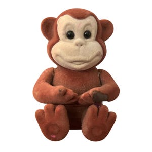 May include: A vintage, seated monkey figurine with a fuzzy, flocked texture. The monkey has a brown body, ears, and feet, with a cream-colored face and belly. It features black eyes and is posed with its hands clasped.