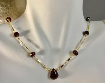 May include: A gold chain necklace with a teardrop-shaped garnet pendant and smaller garnet beads. The necklace is on a white mannequin.