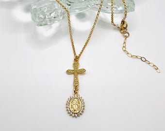 May include: Gold chain necklace with a small cross pendant and a round pendant with a depiction of the Virgin Mary, both adorned with clear cubic zirconia stones.