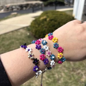 May include: A collection of beaded flower bracelets in shades of pink, blue, white, and purple. The bracelets are linked together and worn on the wrist. The small, delicate flowers have beaded centres.