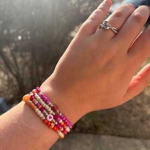 Könnte beinhalten: Ein Stapel aus vier Perlenarmbändern in den Farben Pink, Orange und Weiß. Die Armbänder sind übereinander am Handgelenk getragen und haben einen kleinen rosa Herz-Anhänger.