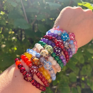 May include: A stack of colourful beaded bracelets with dice charms. Each bracelet features a different colour and type of bead, including red, blue, green, and purple. The dice charms are all different sizes and shapes, and some have numbers on them.