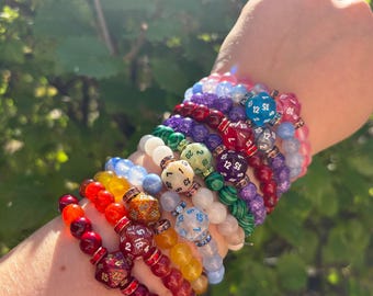 May include: A stack of colorful beaded bracelets with dice charms. Each bracelet features a different color and type of bead, including red, blue, green, and purple. The dice charms are all different sizes and shapes, and some have numbers on them.