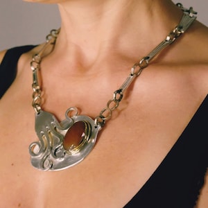 Puede incluir: Un collar de plata con una piedra preciosa grande, ovalada y marr&oacute;n engastada en un bisel de oro. El colgante tiene forma de pulpo con sus tent&aacute;culos enroscados alrededor de la piedra.