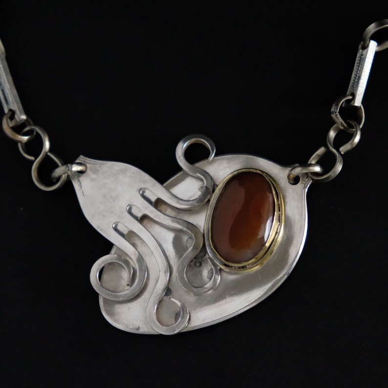 Puede incluir: Collar de plata con una piedra marr&oacute;n engastada en un bisel de oro. El colgante tiene forma de pulpo con tent&aacute;culos rizados.