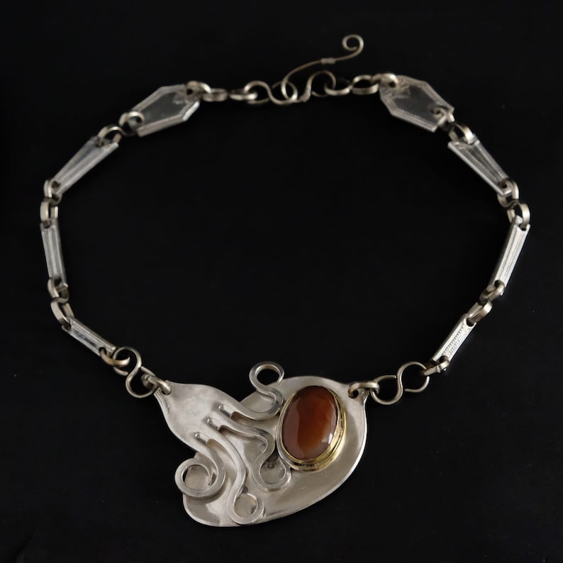 Puede incluir: Un collar de plata con un colgante hecho de una cuchara reutilizada. El colgante presenta una piedra ovalada marr&oacute;n engastada en un bisel dorado y un dise&ntilde;o en espiral.